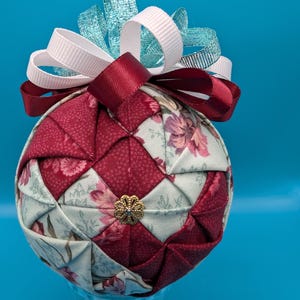 May include: A quilted fabric Christmas ornament with a floral pattern in shades of red, pink, and green. The ornament is topped with a white and blue ribbon bow.