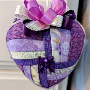 Peut inclure: Ornement en forme de cœur en tissu violet et blanc avec un grand nœud violet et un plus petit nœud violet avec des accents de perles.