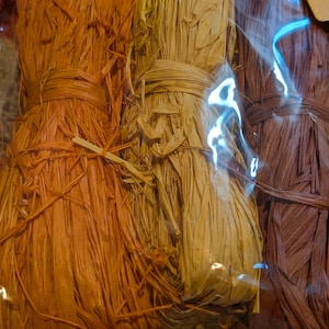 May include: Close-up of several bundles of dried, textured raffia in shades of orange, yellow, and brown, tied with matching raffia. The bundles are wrapped in clear plastic.