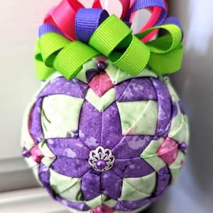 May include: A purple and green fabric Christmas ornament with a pink, green, and purple ribbon bow. The ornament is made of fabric squares sewn together in a quilted pattern.