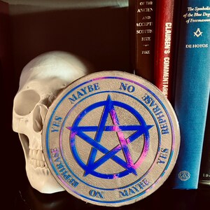 May include: A circular metallic coin featuring a central iridescent blue pentagram. The outer rim is inscribed with the words YES, NO, MAYBE, and REPHRASE repeated around the perimeter. A white human skull model is positioned behind the coin.