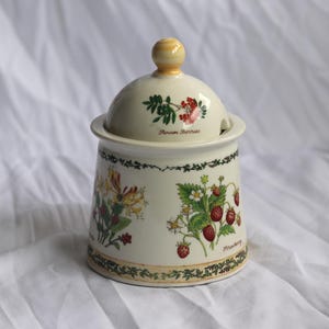 May include: A cream-coloured ceramic sugar bowl with a matching lid featuring a yellow knob. The exterior is decorated with botanical illustrations of strawberries and other plants, including the text "Strawberry" and "Potentilla" in script.