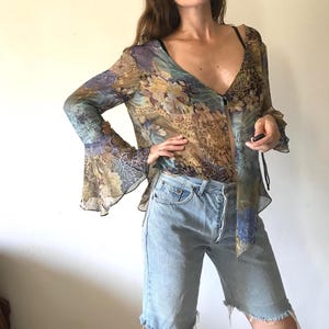 May include: A sheer long-sleeve blouse featuring a multi-colored floral and leopard print pattern with a deep V-neckline and flared bell sleeves, paired with light-wash denim shorts that have a raw, frayed hemline and a button-fly closure.