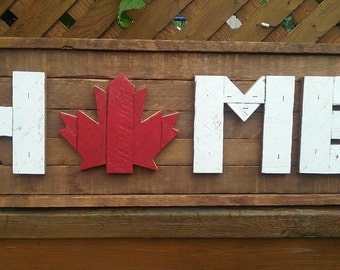 May include: A rectangular wooden sign featuring the word HOME in white block letters, with a red wooden maple leaf replacing the letter O. The sign is constructed from horizontal brown wooden planks with a visible grain and texture throughout.