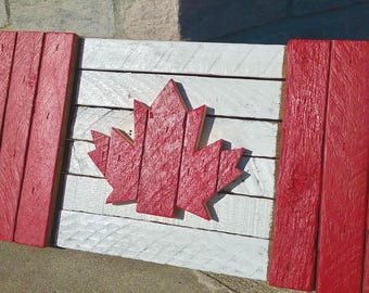 May include: A rectangular wall art piece constructed from horizontal wooden slats painted to resemble the Canadian flag. The design features two vertical red sections on the sides and a central white section with a raised red wooden maple leaf.