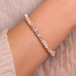 May include: A bracelet composed of a single strand of small, round, off-white beads. Each bead features a distinct pattern of dark brown or black speckles and fine, radiating lines, creating a textured appearance across the entire length of the jewelry.