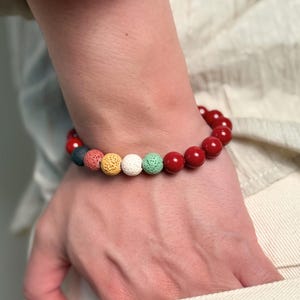 Può includere: Un braccialetto di perline rosse con un mix di perline colorate. Il braccialetto presenta una serie di perline rosse rotonde e lucide e una fila di perline più piccole e strutturate in vari colori, tra cui giallo, verde e blu. Il braccialetto è indossato al polso.
