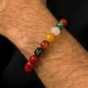 Op de afbeelding: Een armband met kleurrijke, ronde kralen in rood, zwart, geel, wit en groen. Sommige kralen hebben gouden ontwerpen. De armband wordt om een pols gedragen.