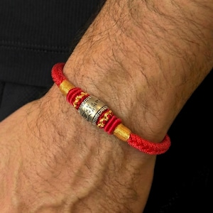 Può includere: Bracciale intrecciato rosso e oro con un elemento centrale cilindrico argentato. Il bracciale è indossato al polso e presenta nodi e dettagli intricati. Il pezzo d'argento ha un testo inciso.