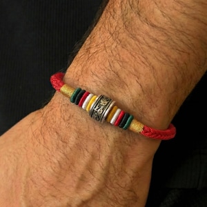 Può includere: Un braccialetto rosso intrecciato con un elemento centrale cilindrico argentato. Il braccialetto presenta accenti colorati in oro, verde, bianco e rosso. Il braccialetto è indossato al polso.