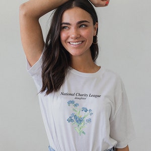 May include: White t-shirt with a blue floral design and the text "National Charity League daughter" printed on the front.