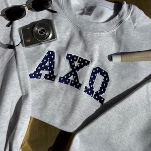 May include: A light gray sweatshirt with the Greek letters ΑΧΩ in navy blue with white polka dots. The sweatshirt is paired with black sunglasses, a silver digital camera, and a lip balm tube with the text "SUMMER FRIDAYS".