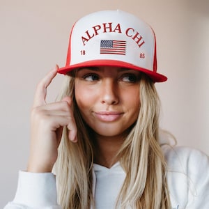 Può includere: Un cappello da baseball bianco e rosso con una parte posteriore in rete. La parte anteriore del cappello ha il testo "ALPHA CHI" in ricamo rosso e una piccola bandiera americana. I numeri "18" e "85" sono ricamati in rosso su entrambi i lati della bandiera.