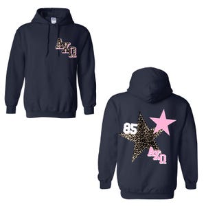 May include: Navy blue hooded sweatshirt with a pink and leopard print star design on the back. The front features the Greek letters &Alpha;&Chi;&Omega; in a leopard print design. The number 85 is also visible.