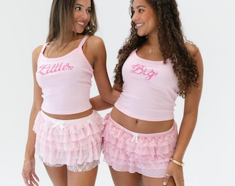May include: Two people wearing pink tank tops with the words "Little" and "Big" embroidered on them. They are also wearing pink ruffled skirts.
