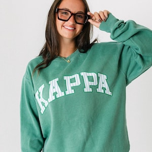 以下が含まれることがあります： 胸に白いブロック文字で「KAPPA」と書かれたグリーンのスウェットシャツ。スウェットシャツは長袖でクルーネックです。文字はストライプのデザインです。