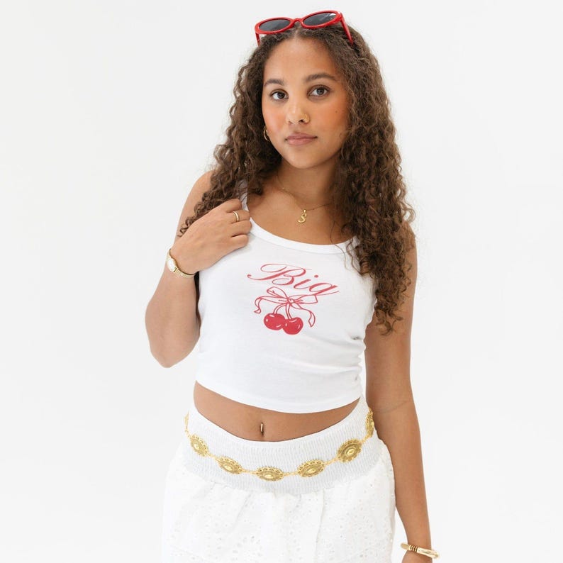 May include: White cropped tank top with a red cherry and bow graphic and the word "Big" in cursive. The model is wearing a gold chain belt and white skirt.