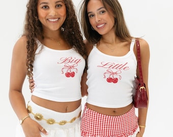 May include: Two women wearing white tank tops with red cherry graphics and the words "Big" and "Little" on them. One woman is wearing a white skirt with gold embellishments and the other is wearing a red and white gingham skirt.