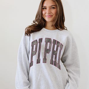 Pode incluir: Um moletom cinza claro com as letras "PI PHI" em um design de estampa de leopardo. As letras são contornadas em uma cor bordô escura. O moletom é de estilo casual.
