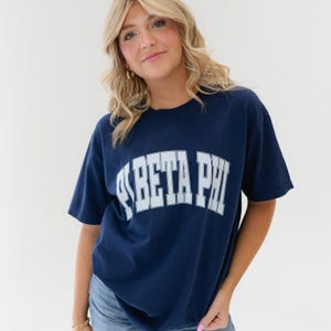 以下が含まれることがあります： 前面に白い「Phi Beta Phi」のグラフィックがプリントされたネイビーブルーのTシャツです。