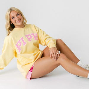 Puede incluir: Una sudadera amarilla clara con las letras rosas "PI PH" en el pecho. La persona lleva pantalones cortos rosas y calcetines blancos con zapatillas. El fondo es blanco.