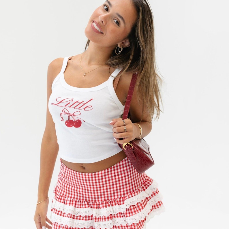 May include: A white tank top with a red cherry graphic and the word "Little" printed on the front. The model is wearing a red and white gingham skirt with white lace trim.