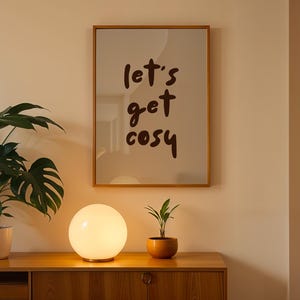 Peut inclure: Un meuble en bois avec une lampe sphérique et une petite plante en pot. Une affiche encadrée sur le mur indique "let's get cosy" en écriture brune. Une grande plante en pot avec des feuilles vertes est à gauche.
