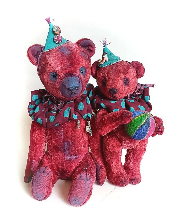 May include: Two red teddy bears wearing colorful hats and a polka dot collar. The larger bear is sitting down, while the smaller bear is holding a colorful ball.