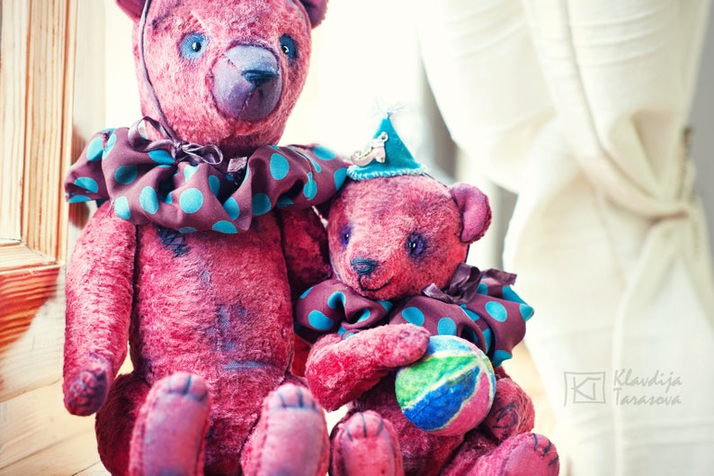 May include: Two plush teddy bears with pink fur, one wearing a brown and teal polka dot collar and the other wearing a teal and brown polka dot collar and a party hat. The smaller bear is holding a colorful ball.
