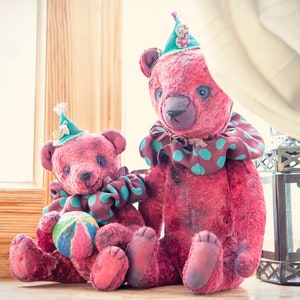 May include: Two pink teddy bears wearing green and brown clown costumes. The larger bear is sitting with its legs crossed, while the smaller bear is holding a colorful ball. Both bears are wearing green and white hats.