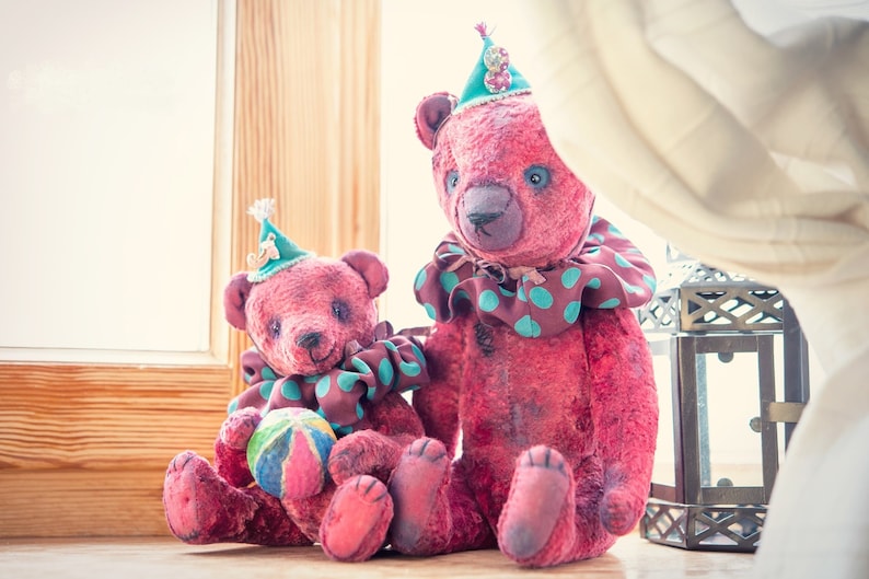 May include: Two pink teddy bears wearing green and brown clown costumes. The larger bear is sitting with its legs crossed, while the smaller bear is holding a colorful ball. Both bears are wearing green and white hats.