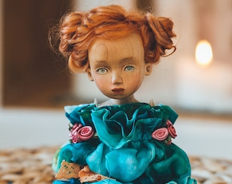 May include: A doll with vibrant ginger hair styled in two buns, featuring a pale face with light green eyes and rosy cheeks. The doll wears a ruffled teal dress adorned with small pink fabric roses and a piece of beige lace at the neckline.