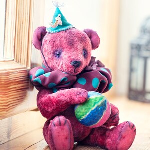 May include: A pink teddy bear wearing a blue and green polka dot scarf and a blue hat with a small bird figurine. The bear is holding a green, blue, and yellow ball.