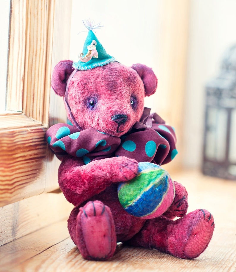 May include: A pink teddy bear wearing a blue and green polka dot scarf and a blue hat with a small bird figurine. The bear is holding a green, blue, and yellow ball.