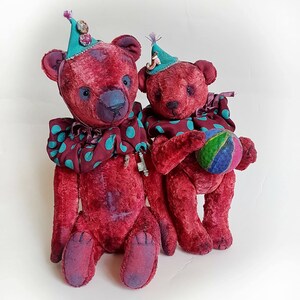 May include: Two red teddy bears wearing colorful clown hats and collars. The larger bear is holding a small ball.