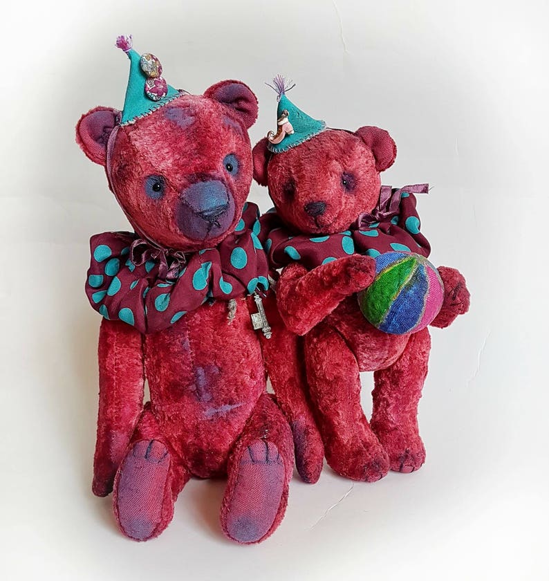 May include: Two red teddy bears wearing colorful clown hats and collars. The larger bear is holding a small ball.