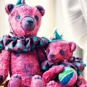 May include: Two pink teddy bears wearing colorful hats and a polka dot collar. The larger bear is sitting with its legs crossed and the smaller bear is sitting with its legs crossed and holding a colorful ball.