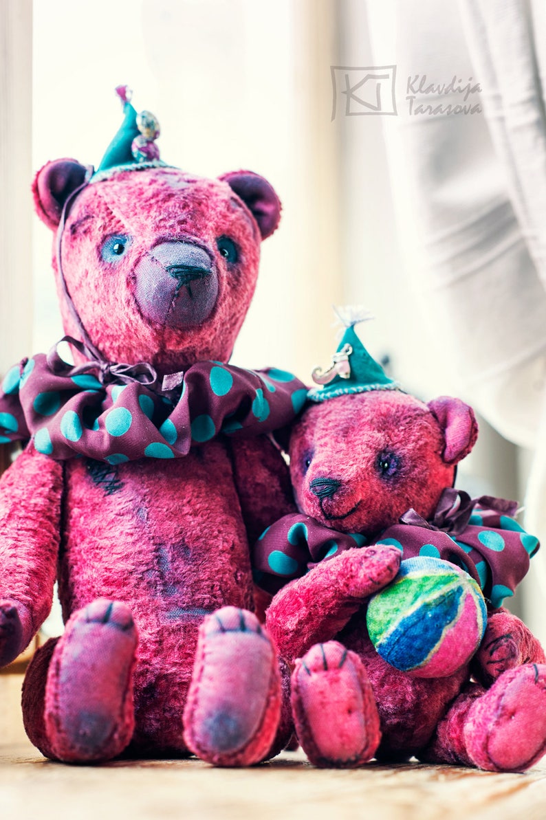 May include: Two pink teddy bears wearing colorful hats and a polka dot collar. The larger bear is sitting with its legs crossed and the smaller bear is sitting with its legs crossed and holding a colorful ball.