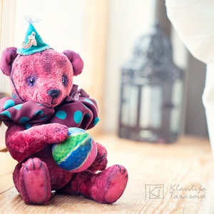 May include: A handmade pink teddy bear wearing a blue and brown polka dot scarf and a blue hat with a small toy on top. The bear is holding a colorful ball in its paws.