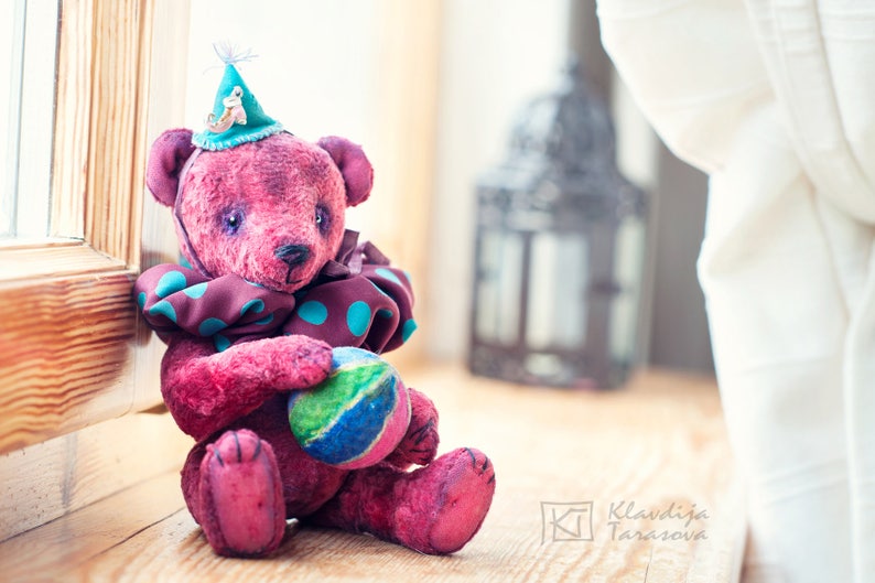 May include: A handmade pink teddy bear wearing a blue and brown polka dot scarf and a blue hat with a small toy on top. The bear is holding a colorful ball in its paws.