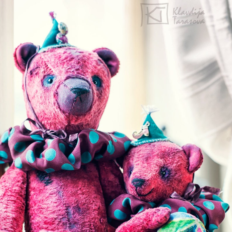 May include: Two pink teddy bears wearing teal and purple hats and matching outfits with teal polka dots. The larger bear is standing and the smaller bear is sitting in its arms. The bears have black eyes and noses.