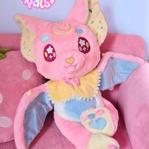 May include: A plush pink bat-like creature with large ears, blue wings, and a yellow and white collar. The plush has star-shaped eyes and a heart-shaped nose. The text "Sugarbell Pals" is above the plush.