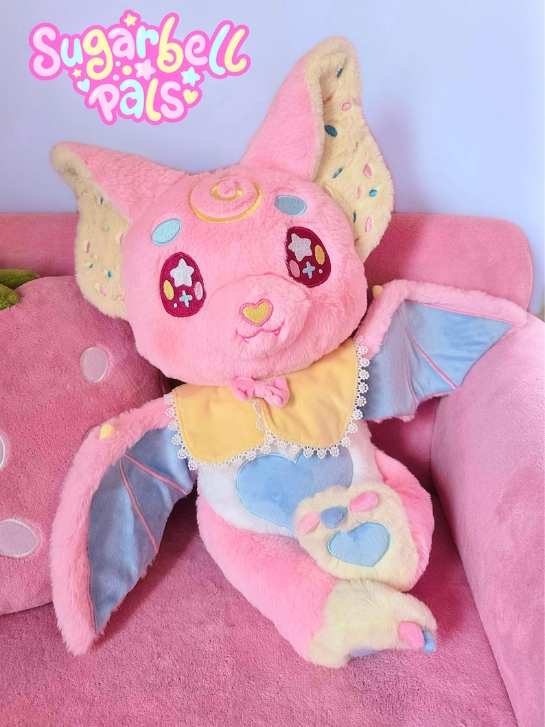 May include: A plush pink bat-like creature with large ears, blue wings, and a yellow and white collar. The plush has star-shaped eyes and a heart-shaped nose. The text "Sugarbell Pals" is above the plush.