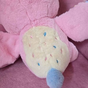 May include: A large, plush pink stuffed animal with a light yellow belly. The belly is decorated with small blue and pink accents. The tail is a fluffy blue. The toy is lying on a pink surface.