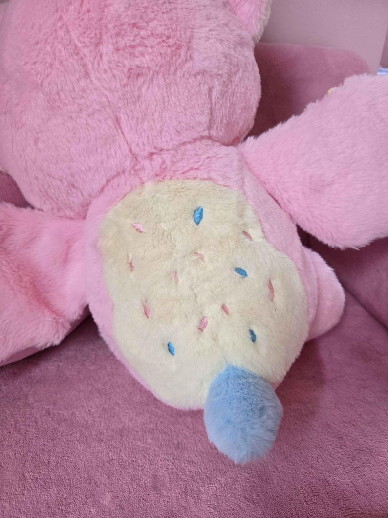 May include: A large, plush pink stuffed animal with a light yellow belly. The belly is decorated with small blue and pink accents. The tail is a fluffy blue. The toy is lying on a pink surface.