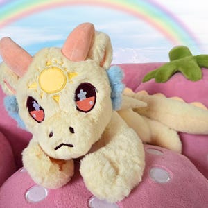 Puede incluir: Un peluche de dragón crema con un emblema de sol en la frente, descansando sobre un cojín rosa con forma de hongo. El dragón tiene cuernos rosas, detalles azules en las orejas y ojos grandes y expresivos. Un peluche verde en forma de estrella está cerca, con un arcoíris de fondo.