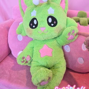 May include: A plush green creature with large eyes, star accents, and a pink star on its chest. The creature has large ears, paw pads, and the text "Sugarbell Pals" in a playful font.