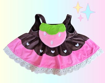 Puede incluir: Vestido para mascotas rosa y marrón con diseño de fresa. El vestido presenta un frente en forma de fresa rosa con una parte superior de hoja verde y detalles de corazón blanco. La falda tiene un diseño en capas rosa y marrón con un ribete de encaje blanco.