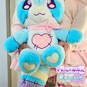 Könnte beinhalten: Eine Plüsch-Patchwork-Waschbär-Puppe in Pastellblau, Lila und Gelb. Die Puppe hat große, lila umrandete Augen, ein rosa Herz auf der Brust und eine passende Schleife. Der Text "Patchwork Raccoon" und "Sugarbell Pals" sind sichtbar.