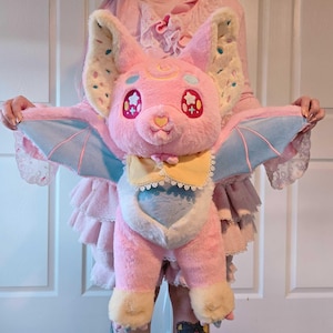 May include: A plush pink bat with large ears, star-shaped eyes, and light blue wings. The bat has a yellow bow tie and a heart-shaped nose. The wings are light blue with pink accents. The ears are cream-colored with colorful sprinkles.